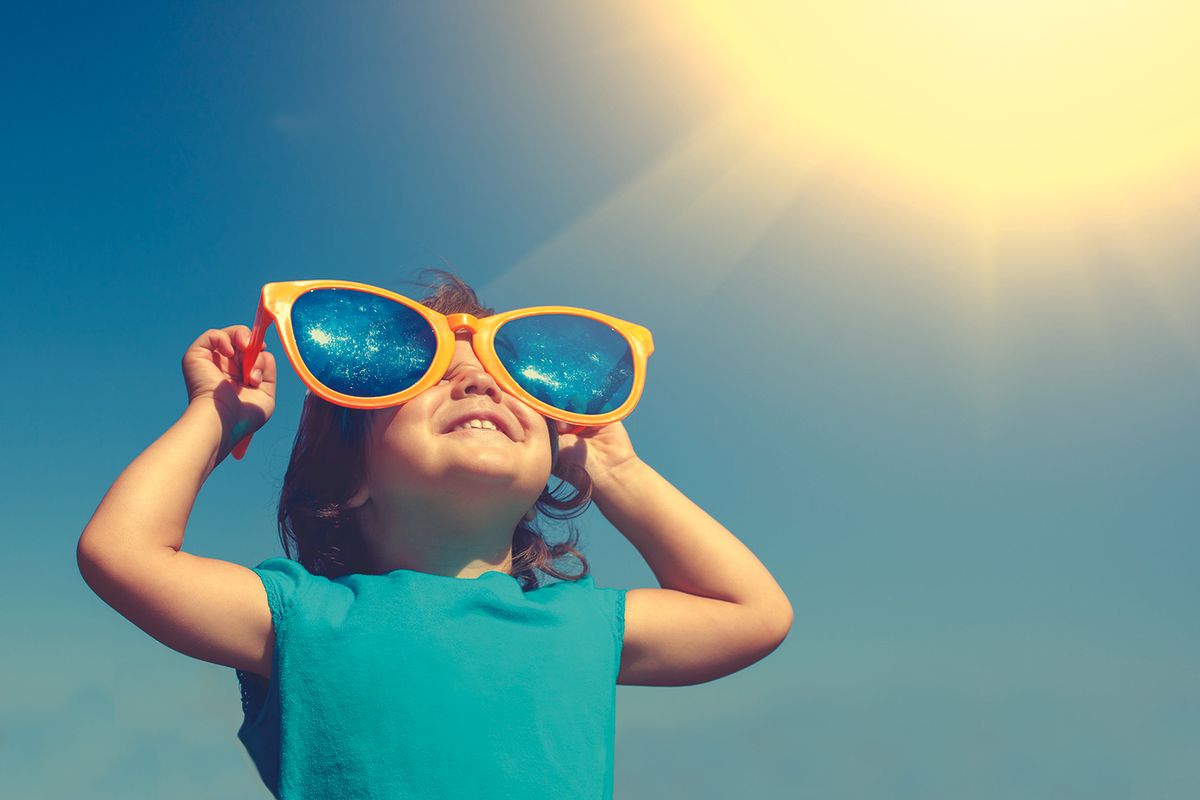 Young girl wearing bright orange oversized sunglasses, looking up at the sunny sky.