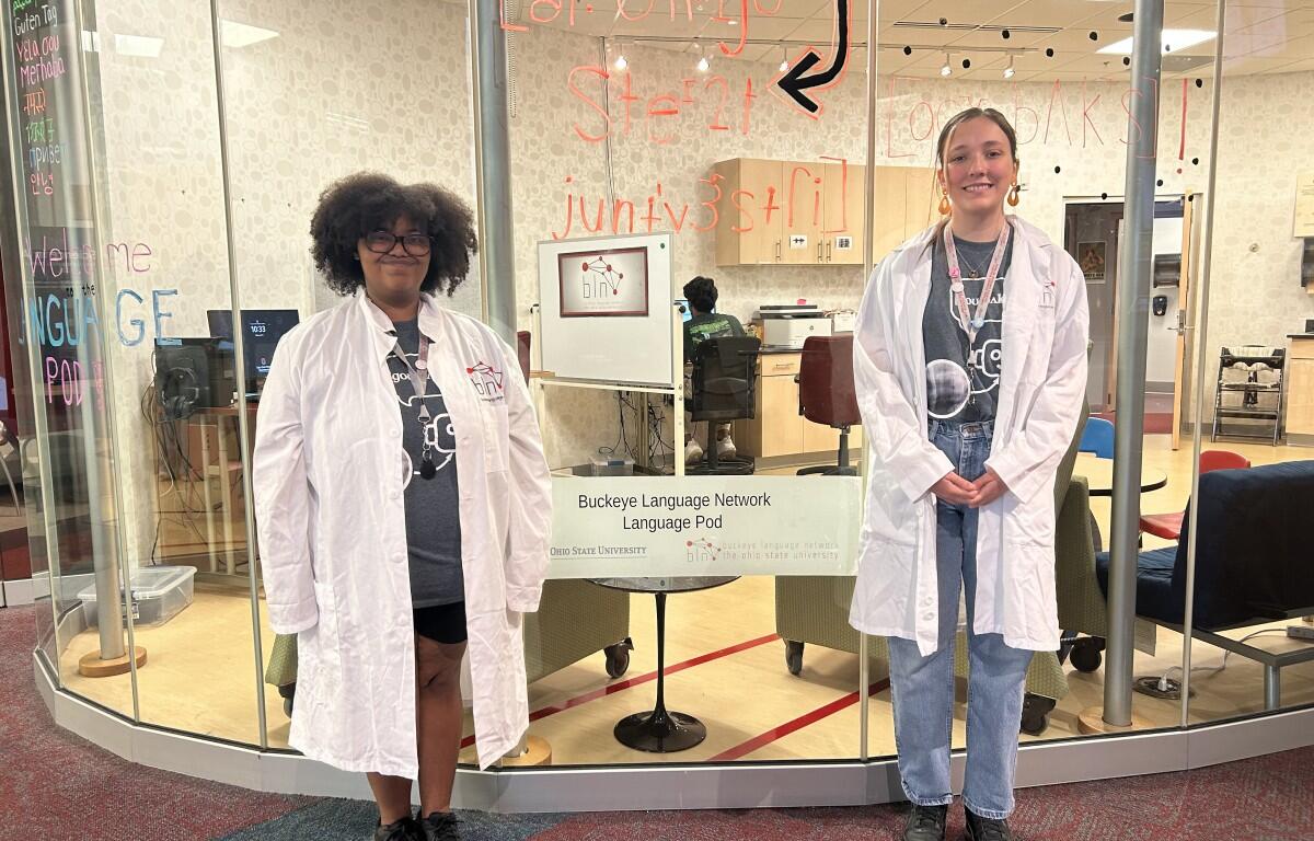 Two students in white lab coats stand outside a glass-walled Buckeye Language Network Language Pod; OSU signage visible inside.
