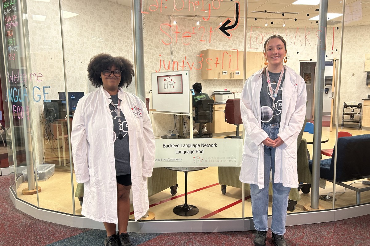 Two students in white lab coats stand outside a glass-walled Buckeye Language Network Language Pod; OSU signage visible inside.