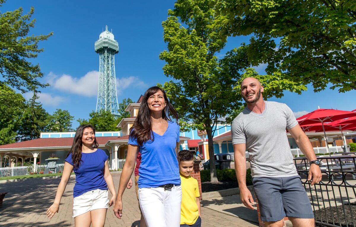 Family of four walking together in a sunny park with a tall lattice observation tower in the background.