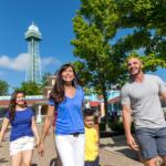 Family of four walking together in a sunny park with a tall lattice observation tower in the background.