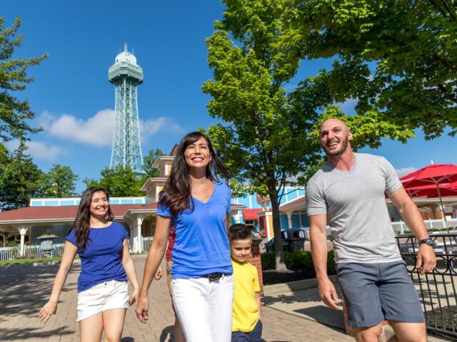 Family of four walking together in a sunny park with a tall lattice observation tower in the background.