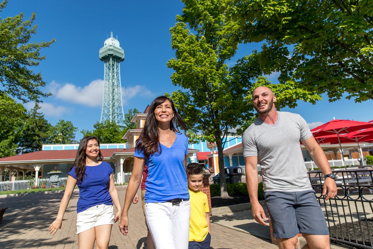 Family of four walking together in a sunny park with a tall lattice observation tower in the background.