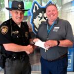 Sheriff in uniform shakes hands with a smiling man in a gray polo, handing over a white envelope, in front of a blue wolf logo backdrop.
