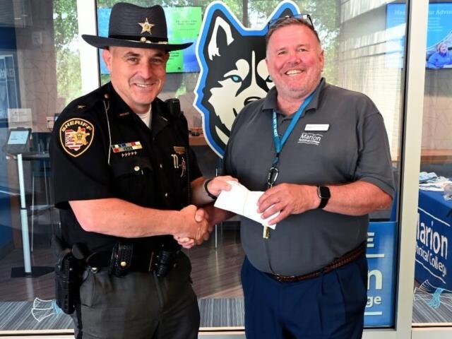Sheriff in uniform shakes hands with a smiling man in a gray polo, handing over a white envelope, in front of a blue wolf logo backdrop.