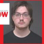 Mugshot-style photo of a young man with glasses and dark hair wearing a black shirt, against a gray backdrop, with a Marion County NOW logo overlay on the left and a red background.