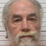 Mugshot of an elderly man with gray hair and a full beard, wearing an orange jumpsuit, standing against a light striped backdrop.