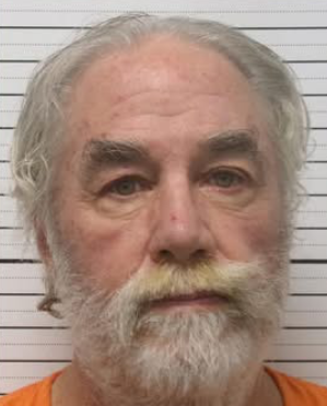 Mugshot of an elderly man with gray hair and a full beard, wearing an orange jumpsuit, standing against a light striped backdrop.