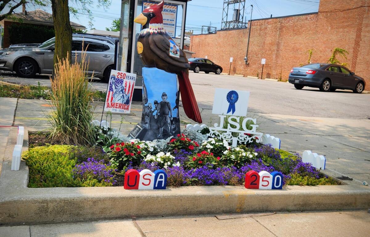 Colorful outdoor memorial with a large carved bird statue surrounded by flowers, banners reading 'USA 250' and '250 Years', and patriotic imagery in a parking lot garden.