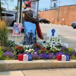 Colorful outdoor memorial with a large carved bird statue surrounded by flowers, banners reading 'USA 250' and '250 Years', and patriotic imagery in a parking lot garden.