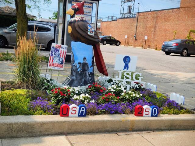 Colorful outdoor memorial with a large carved bird statue surrounded by flowers, banners reading 'USA 250' and '250 Years', and patriotic imagery in a parking lot garden.