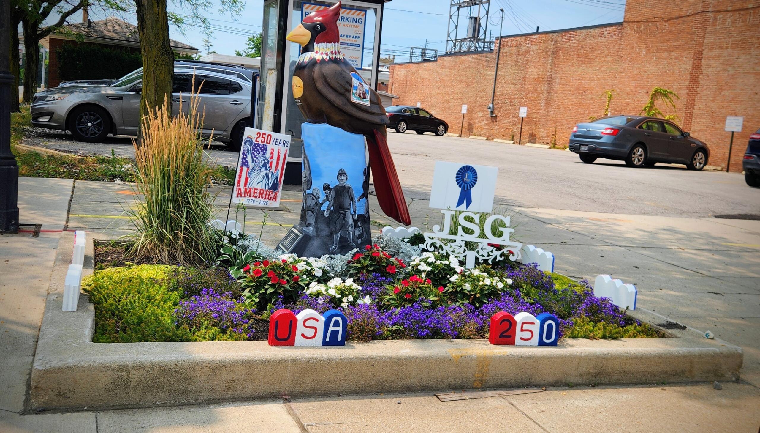 Colorful outdoor memorial with a large carved bird statue surrounded by flowers, banners reading 'USA 250' and '250 Years', and patriotic imagery in a parking lot garden.