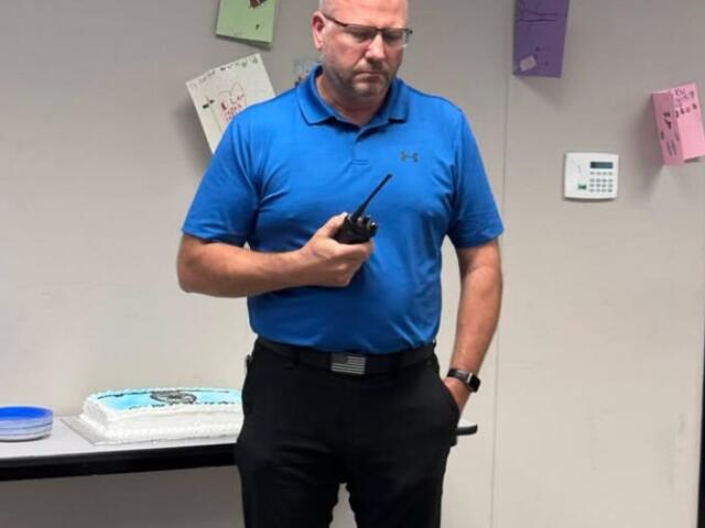 Man in a bright blue polo, wearing safety glasses, holds a radio while standing near a cake on a table in a room with a clock on the wall.