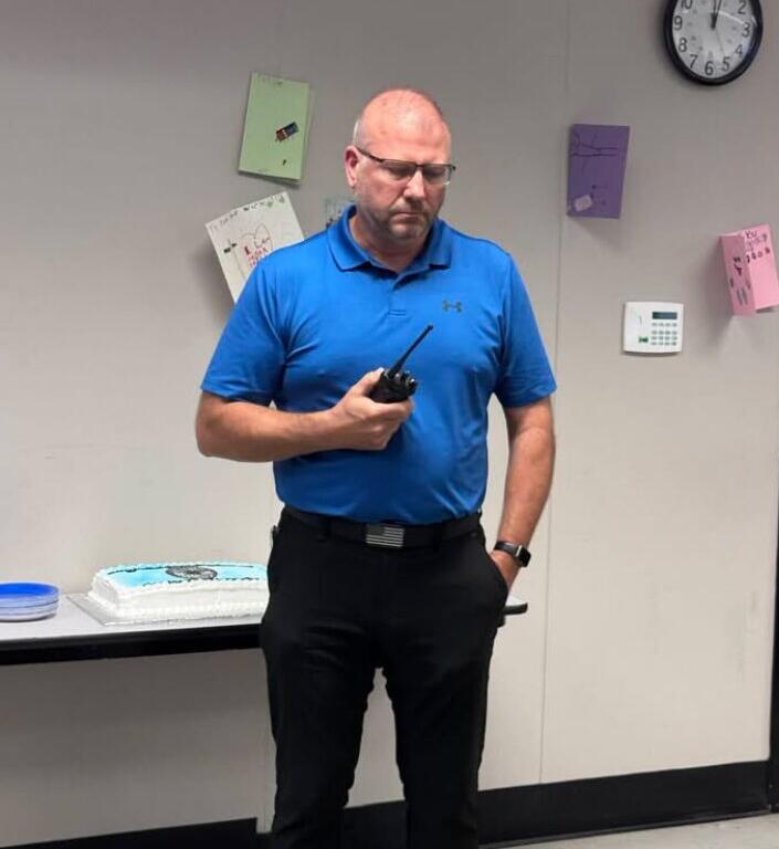 Man in a bright blue polo, wearing safety glasses, holds a radio while standing near a cake on a table in a room with a clock on the wall.