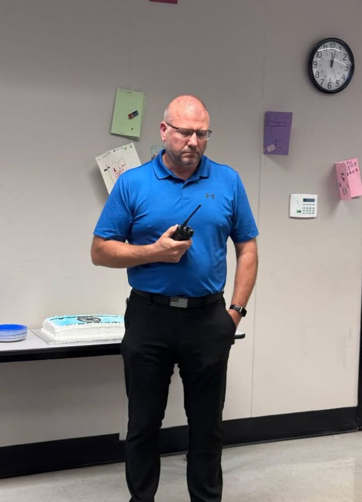 Man in a bright blue polo, wearing safety glasses, holds a radio while standing near a cake on a table in a room with a clock on the wall.