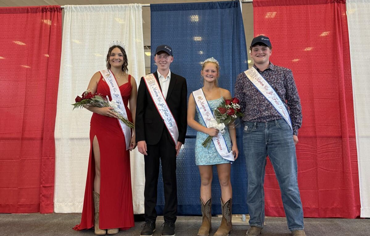 Four pageant contestants stand on a stage in front of red, white, and blue curtains, wearing sashes and crowns, and holding bouquets.