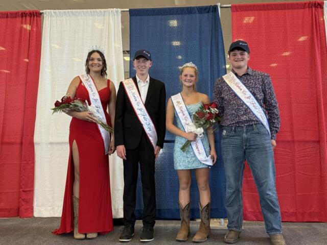 Four pageant contestants stand on a stage in front of red, white, and blue curtains, wearing sashes and crowns, and holding bouquets.