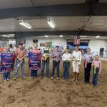 Group of people standing in a livestock arena holding colorful prize ribbons at a county fair award ceremony.