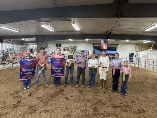 Group of people standing in a livestock arena holding colorful prize ribbons at a county fair award ceremony.