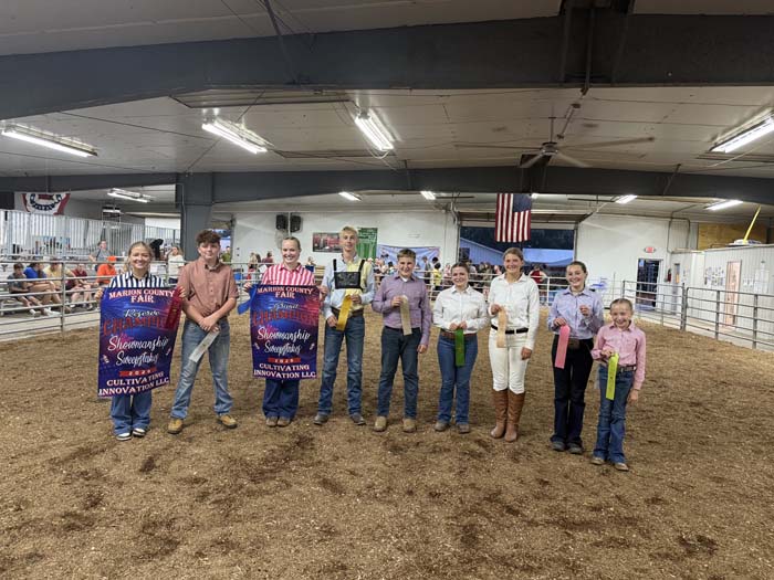 Group of people standing in a livestock arena holding colorful prize ribbons at a county fair award ceremony.