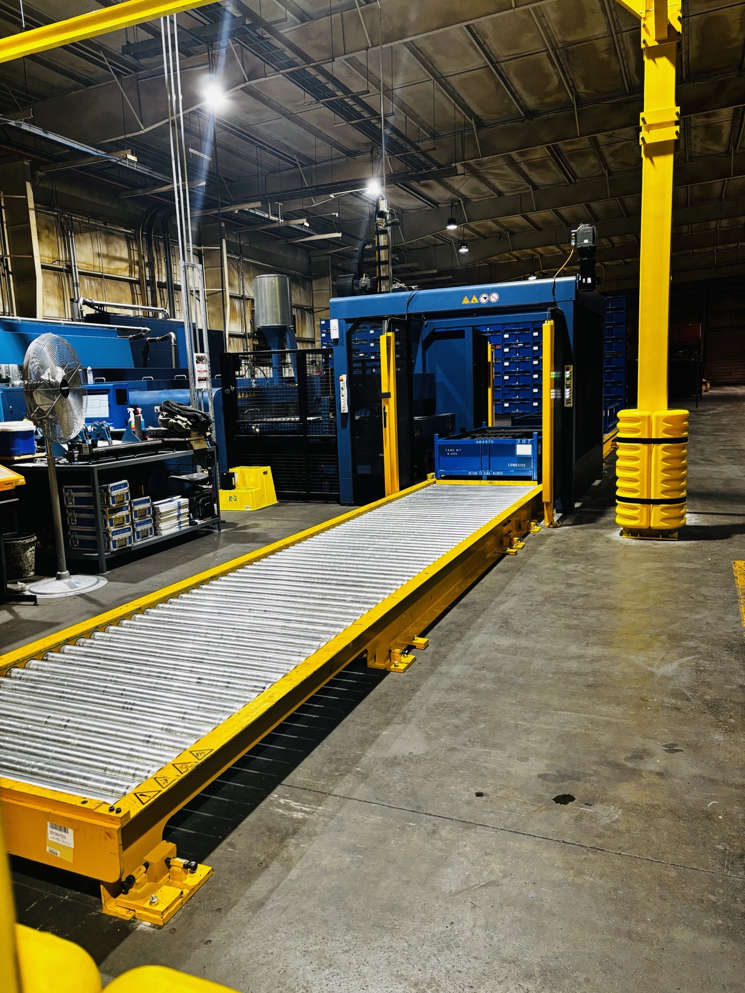 Industrial factory scene with yellow safety guard rails along a roller conveyor and heavy blue machinery in a dimly lit warehouse.