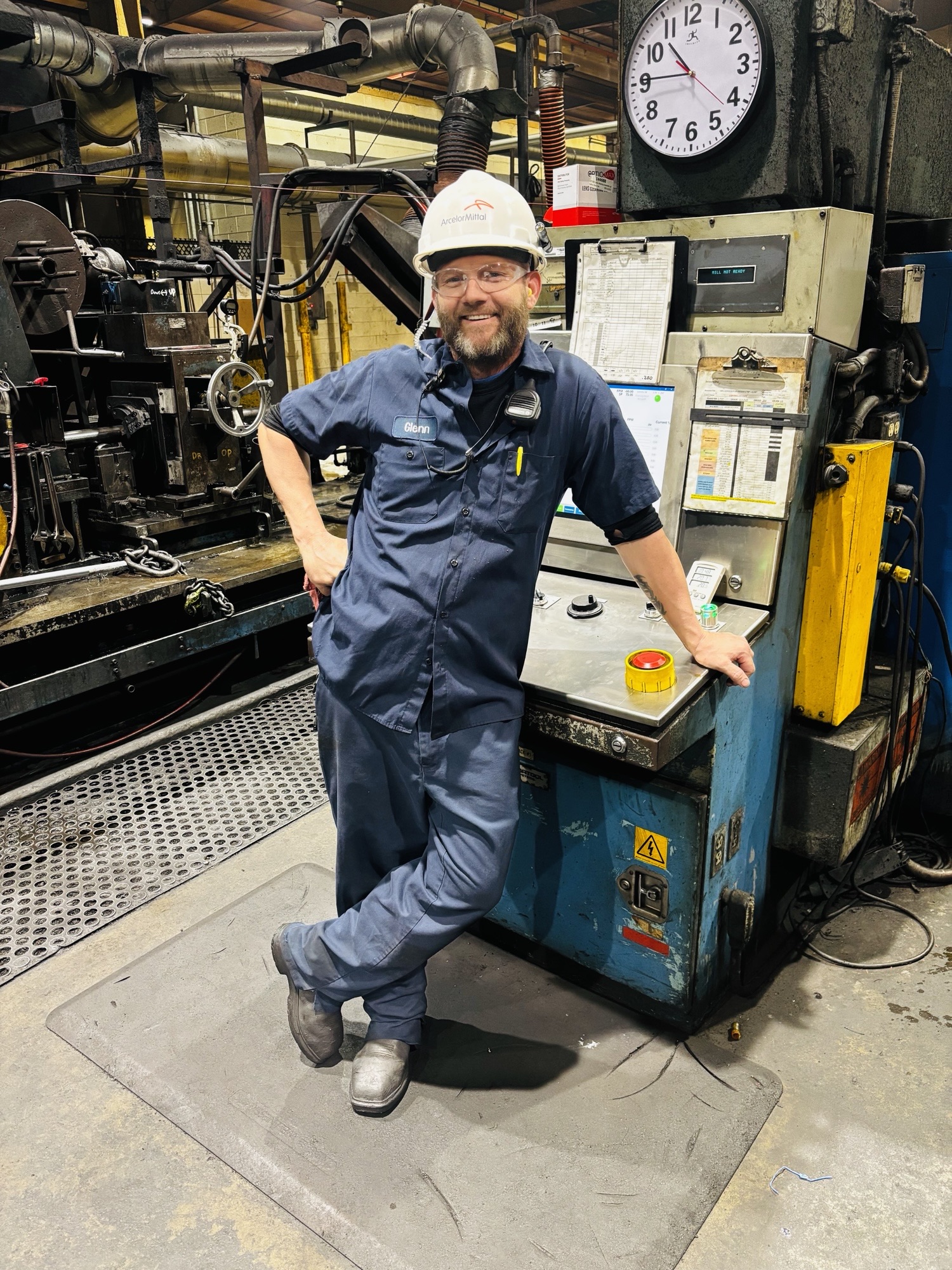 Technician Glenn in blue coveralls and a white hard hat stands beside heavy machinery, smiling.