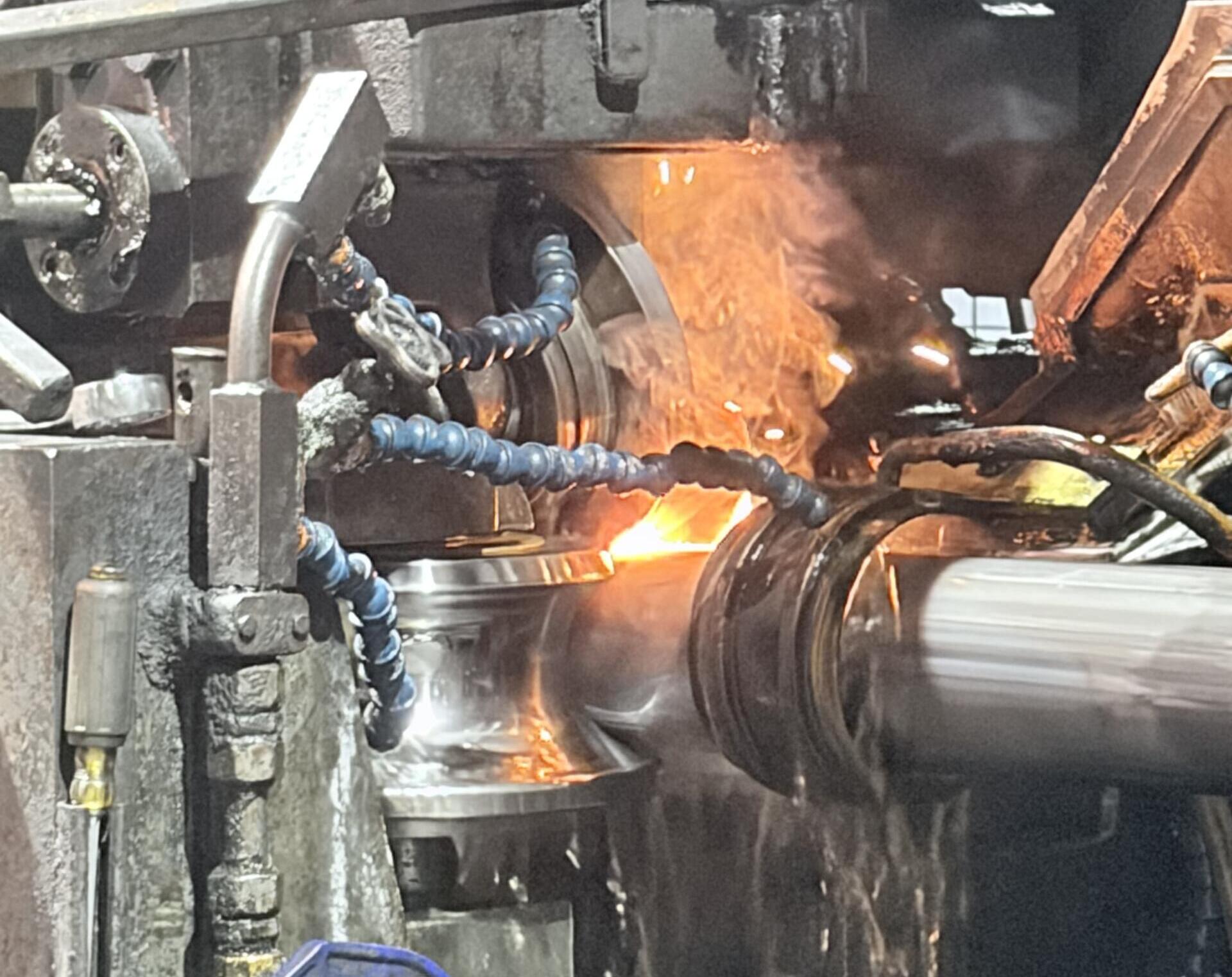 Close-up of a metalworking forge: molten metal glowing as a workpiece is processed, with sparks and industrial machinery around it