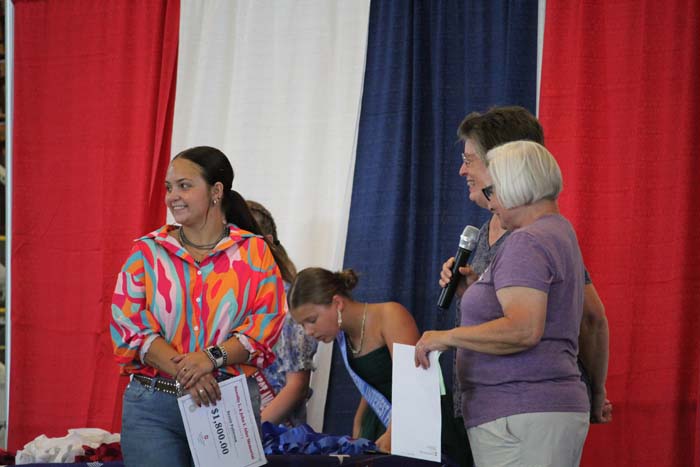 Group of people on a stage with a red, white, and blue backdrop; a smiling woman in a colorful blouse holds a certificate.