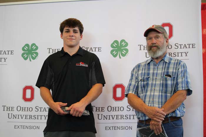 Two men pose for a photo in front of an Ohio State University Extension backdrop; left is a younger man in a black polo, right an older man in a blue plaid shirt and cap?