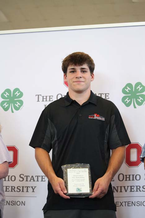 Young man in a black polo shirt holds a plaque at a 4-H/OSU Extension event backdrop curious smile in front of logos and banners from Ohio State University Extension and 4-H clovers.