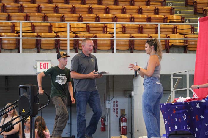Man in a gray shirt hands a plaque to a woman in jeans on a stage inside a gym, with a boy watching nearby.