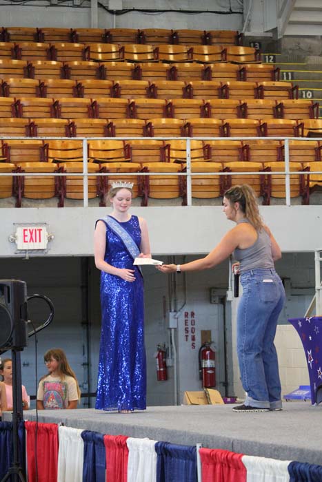 Young pageant contestant in a blue sequin gown and crown receives a certificate from a presenter on a stage, with empty bleachers and red‑white‑blue decorations in the background.