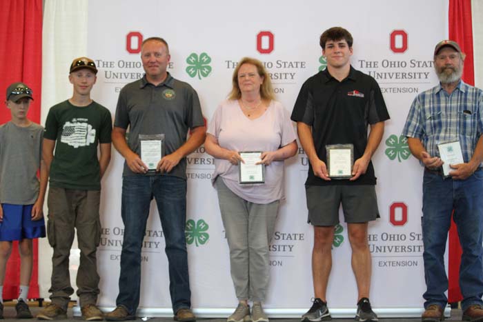 Six people stand in a row on a stage holding certificates, with an Ohio State University backdrop featuring OSU logos and four-leaf clover banners.]