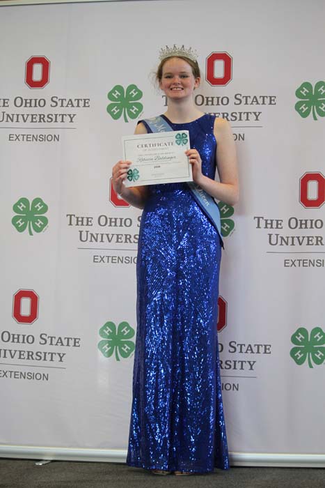 Young woman in a blue sequined gown and crown holds a certificate onstage, with an Ohio State University Extension backdrop.