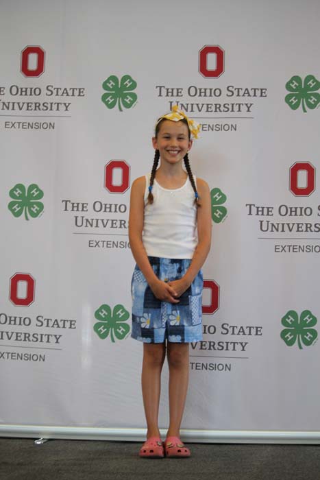 Smiling girl with braided hair and a yellow bow posing in front of an OSU Extension and 4-H backdrop, wearing a white tank top, blue patterned shorts, and pink sandals.