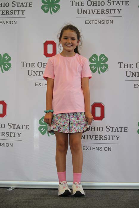 Smiling girl in a pink T-shirt and floral shorts stands in front of a backdrop with The Ohio State University Extension logos.