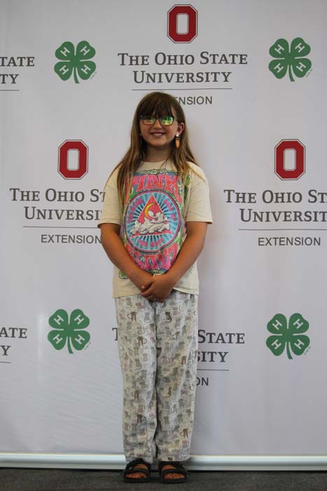 Girl posing for a photo in front of The Ohio State University Extension banner, wearing a unicorn T‑shirt, light patterned pants, sandals, and sunglasses.