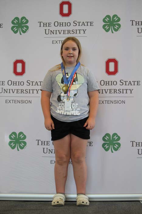 Young girl standing in front of the OSU Extension backdrop, wearing a gray graphic t-shirt, black shorts, white sandals, with medals around her neck.