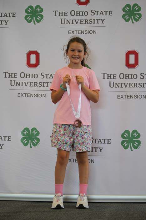 Young girl in a pink shirt and floral shorts stands on a stage, proudly showing a medal on a ribbon with an OSU Extension backdrop behind her. (Informative)