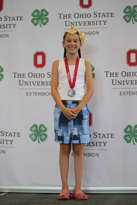 Young girl with braided hair and a yellow bow, wearing a white tank top and blue shorts, standing with a silver medal on a red ribbon in front of an Ohio State University Extension backdrop.