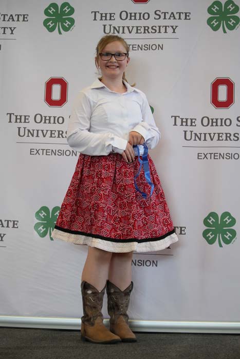Young girl in a white blouse and red patterned skirt posing with a blue ribbon in front of an OSU Extension backdrop.