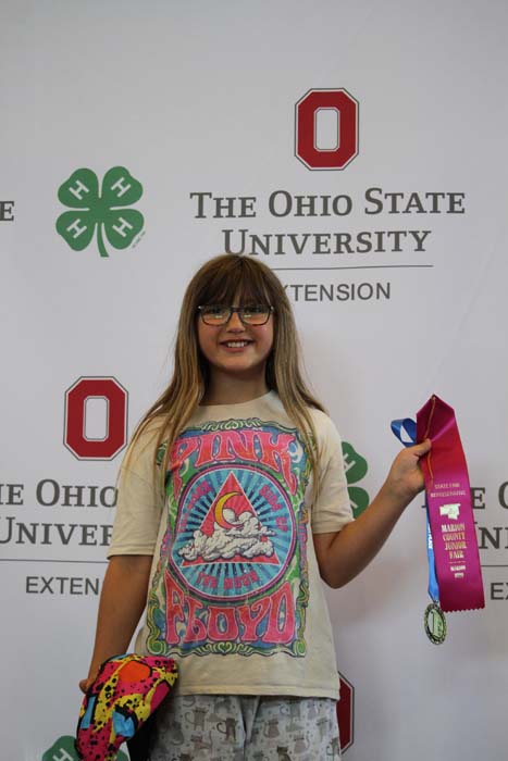 Young girl with glasses and long hair stands in front of an OSU Extension backdrop, proudly holding a pink ribbon prize in one hand.