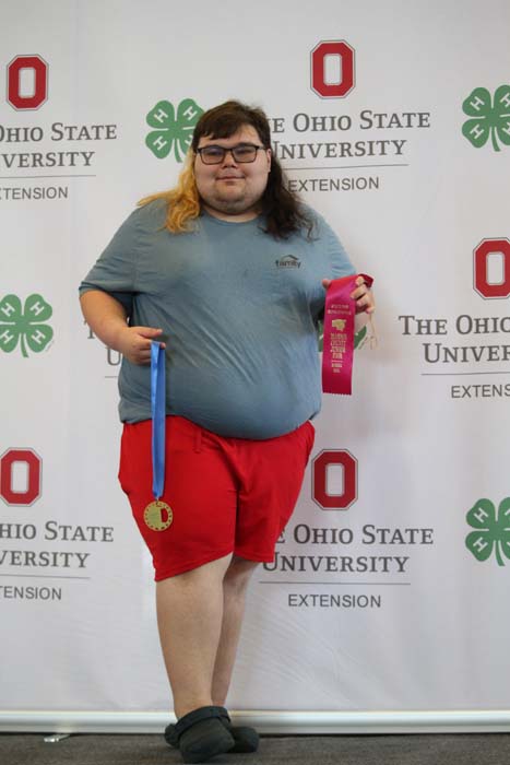 Person wearing a blue shirt and red shorts stands at a ceremony, holding a blue ribbon medal and a pink award ribbon in front of an Ohio State University Extension backdrop.