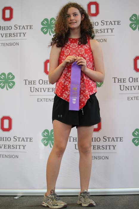 Young girl in a red sleeveless top and black shorts stands on stage, holding a purple award ribbon in front of a backdrop with Ohio State University Extension logos.