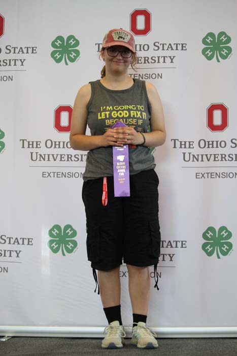 Young woman in a gray tank top and cap stands before an Ohio State University Extension backdrop, holding a purple ribbon.
