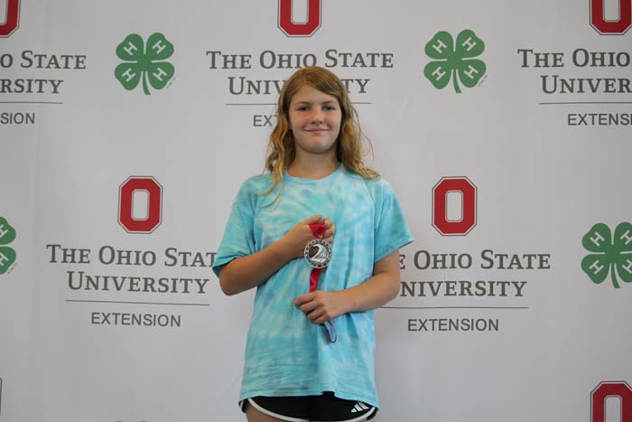 Young girl in a blue tie-dye shirt holding a silver medal with a red ribbon, posed in front of an OSU Extension backdrop.