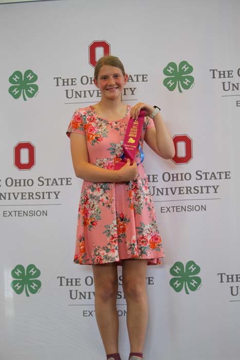 Young woman in a pink floral dress holds a purple award ribbon on a backdrop featuring The Ohio State University Extension and 4-H logos, smiling at the camera.