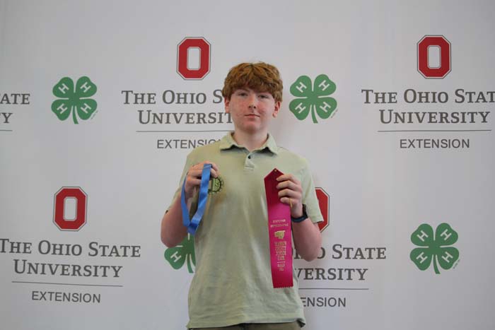 Young person stands in front of an Ohio State University Extension backdrop, holding a blue ribbon and a red ribbon as awards.