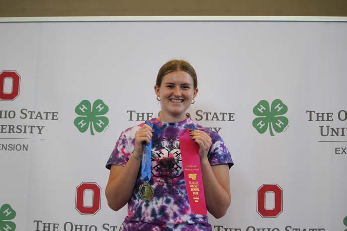 Smiling young woman in a tie-dye shirt holds a gold medal and a pink first-place ribbon in front of an Ohio State backdrop with logos.