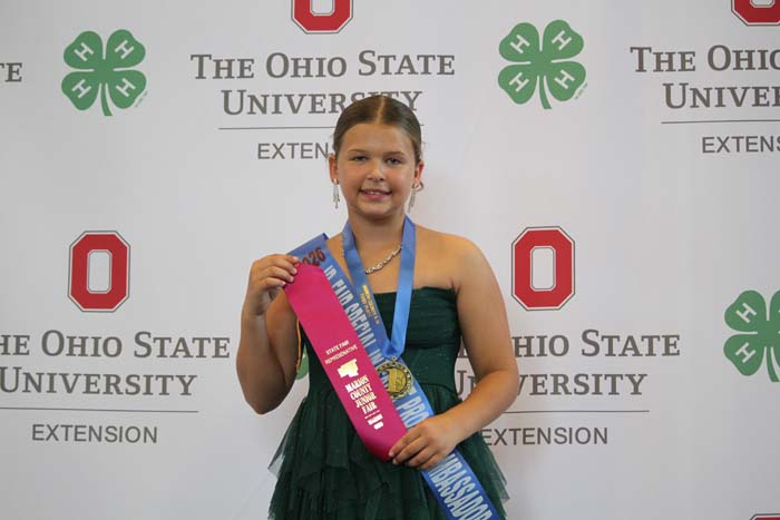 Young girl in a green dress wears a gold medal and pink and blue sashes, posing in front of an Ohio State University backdrop.